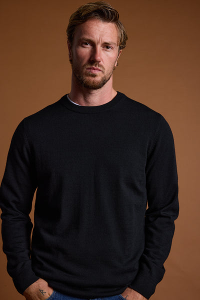 A man with light brown hair and a beard, wearing a black Barkers Skyland ZQrx Merino Crew sweater made from sustainable merino wool, stands against a plain brown background with a neutral expression and his hands in his pockets.