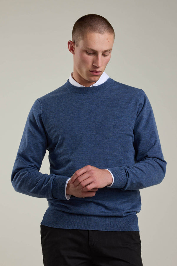 A person wearing a white collared shirt under a Barkers Skyland ZQrx Merino Crew blue sweater and black pants stands against a plain background, looking down with hands clasped in front.