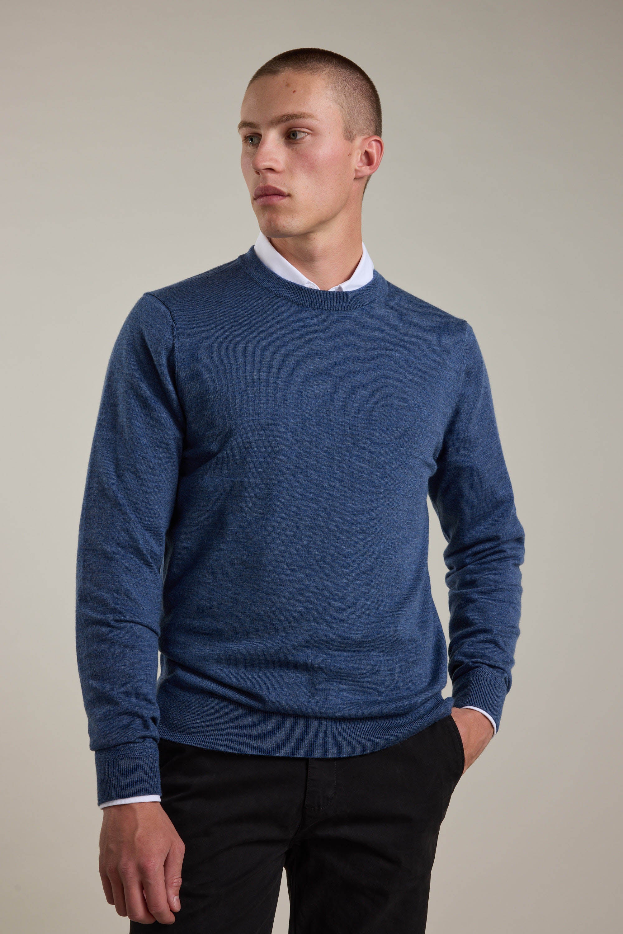 A man in a blue Barkers Skyland ZQrx Merino Crew, made from ethically sourced New Zealand merino wool, stands with one hand in his pocket and looks to the side against a plain light background.