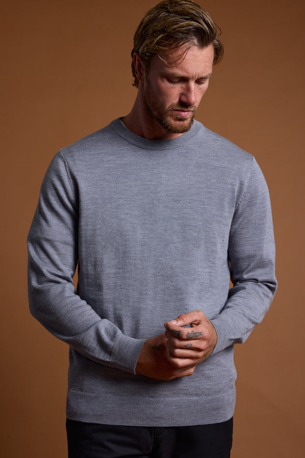 A man with light brown hair and beard wears the Barkers Skyland ZQrx Merino Crew sweater in light gray, standing against a brown background, looking down with hands clasped in front.
