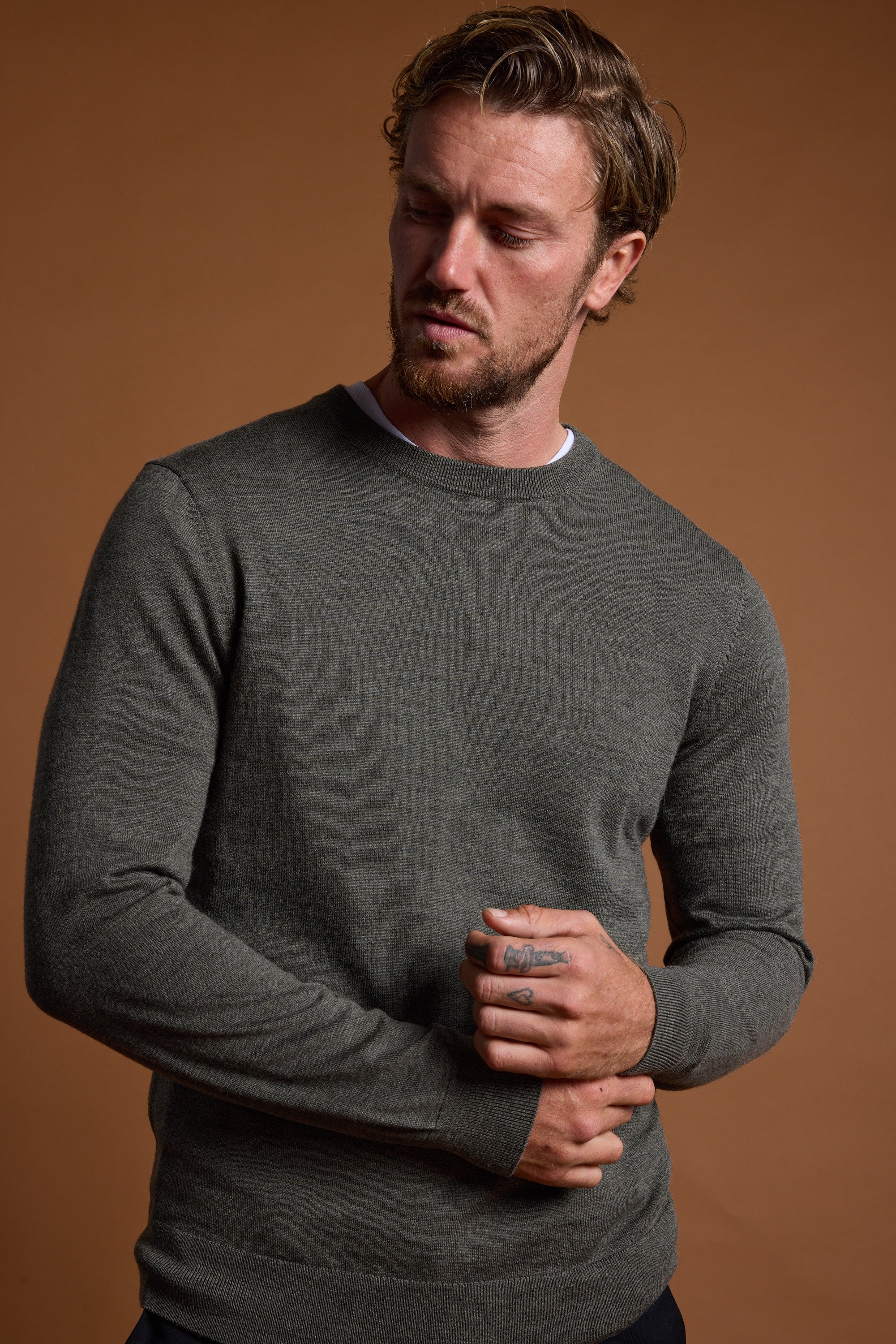A man with wavy hair and a short beard stands against a brown background, wearing the Barkers Skyland ZQrx Merino Crew sweater. He looks down to his right while holding one hand in the other.