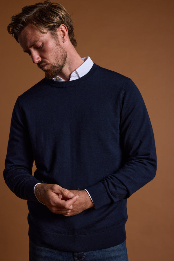 A man with light brown hair and a beard wears the Barkers Skyland ZQrx Merino Crew in navy over a white collared shirt and blue jeans, standing against a brown background with his hands loosely clasped, looking down.