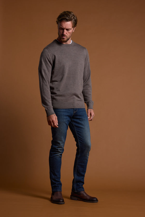 A man stands against a brown background, wearing the Barkers Skyland ZQrx Merino Crew sweater in grey, blue jeans, and brown shoes. He looks slightly downward with one hand by his side and the other touching his thigh.