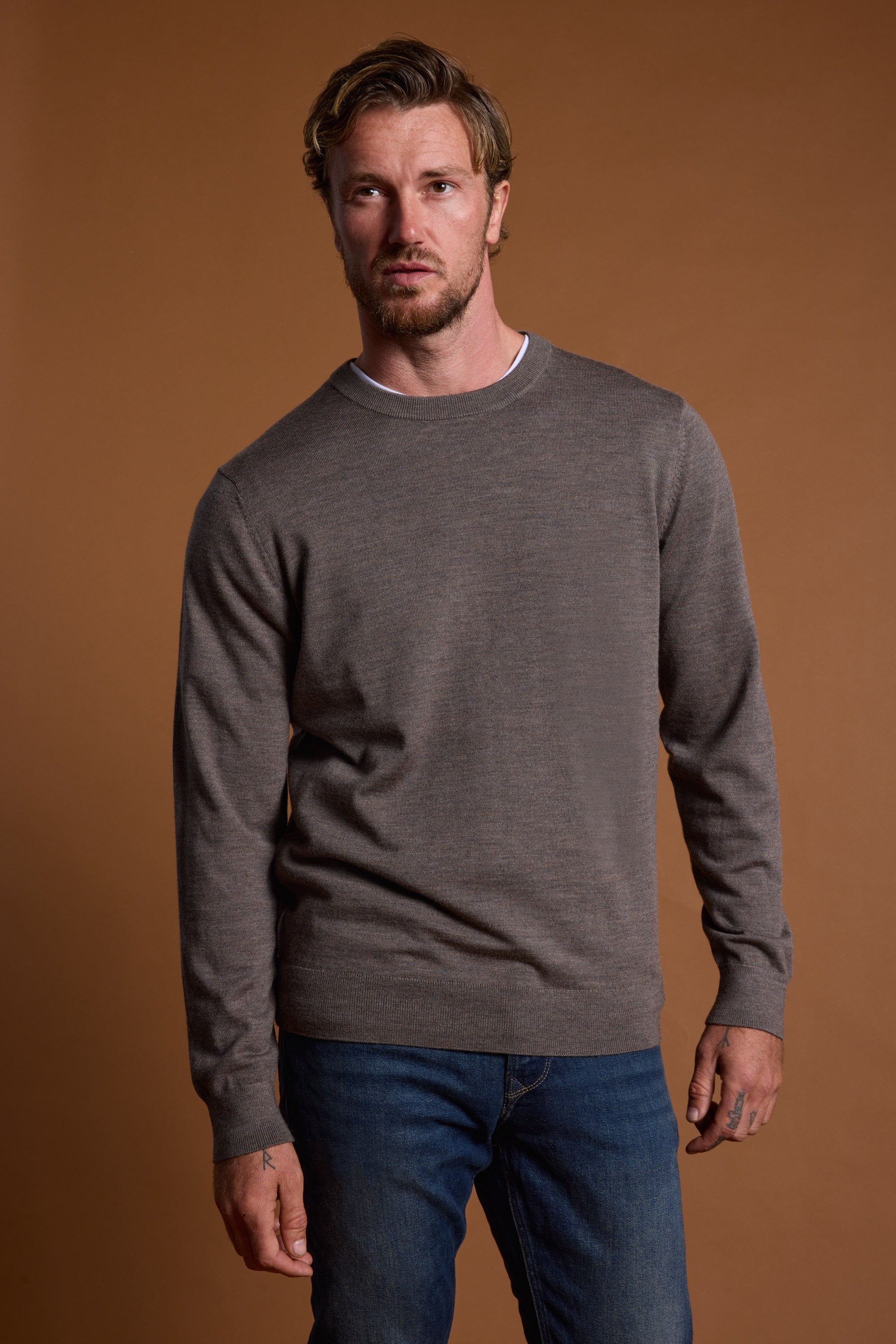 A man with short, wavy hair and facial hair stands against a plain brown background, wearing the Barkers Skyland ZQrx Merino Crew sweater in brownish-gray over a white shirt and blue jeans, looking slightly to the side with a neutral expression.
