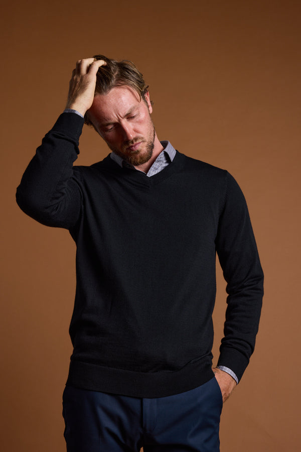 A man in a Barkers Skyland ZQrx Merino Vee sweater and blue pants stands against a brown background, eyes closed, one hand on his head and the other in his pocket, appearing thoughtful or stressed.