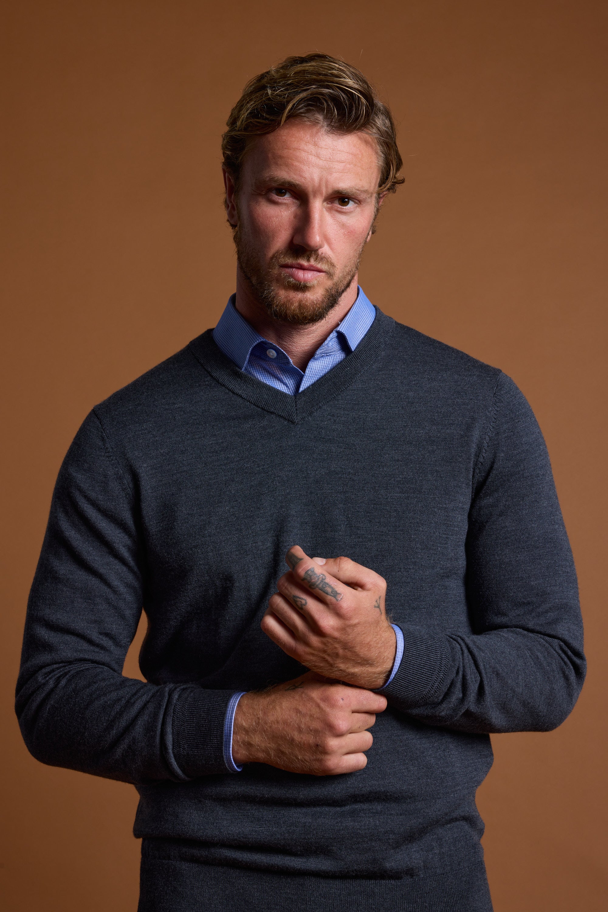 A man with short, wavy hair and a trimmed beard wears the Barkers Skyland ZQrx Merino Vee dark gray sweater over a blue collared shirt, standing against a brown background and looking seriously at the camera.
