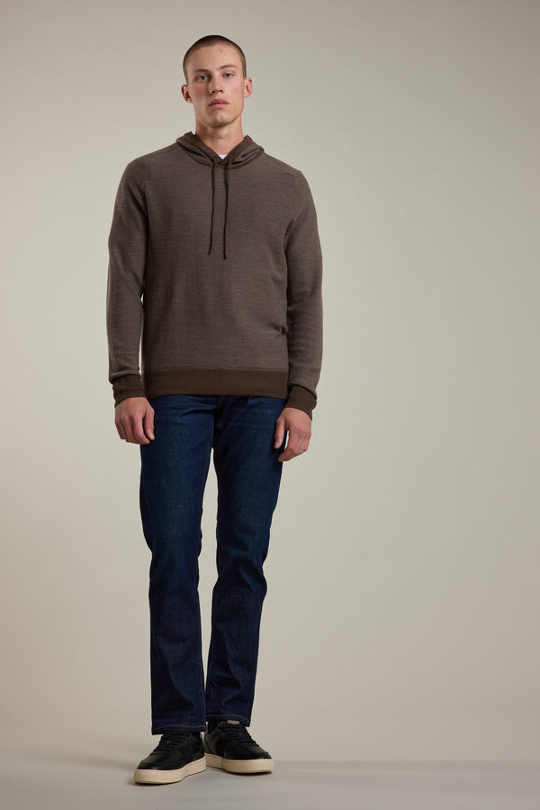 A young man with short hair stands against a plain background, wearing the Barkers Roxbury Merino Hoody in brown, blue jeans, and black sneakers. This lightweight sweater is made from 100% merino wool. His arms are relaxed at his sides.