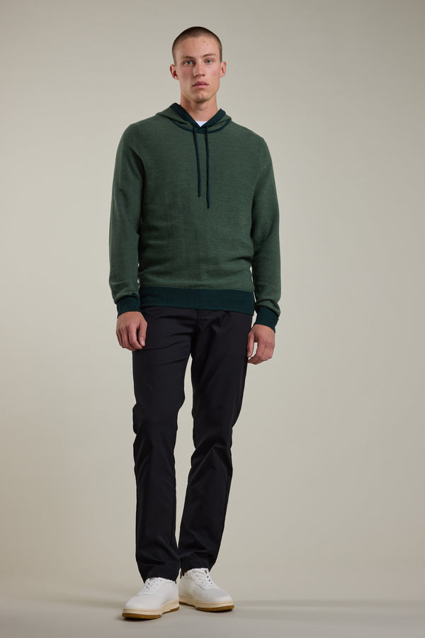 A person with short hair and a neutral expression wears the Barkers Roxbury Merino Hoody in dark green (100% merino wool), paired with black pants and white sneakers, standing against a plain background.