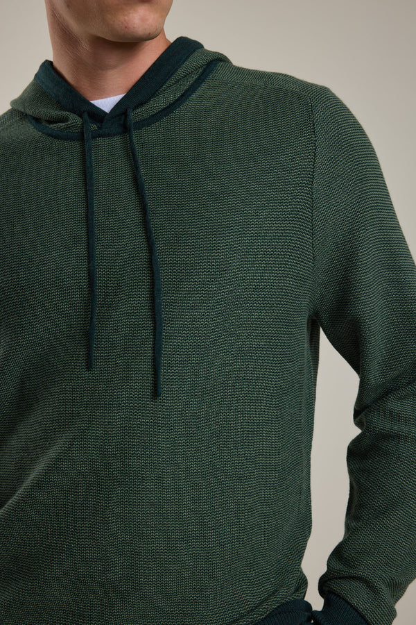 A person wears the Barkers Roxbury Merino Hoody in textured dark green, made from 100% merino wool, shown from shoulders to waist against a neutral background. The face is partially out of frame.