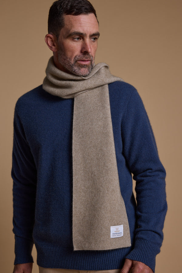 A man with short dark hair and a beard wears a blue sweater and the NZ Made Perino Scarf by Woolyarns Perino, crafted from a merino possum silk blend, against a plain tan background. He looks to his left with one hand at his side.