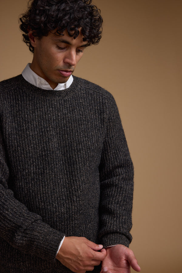 A person with curly dark hair wears the Barkers Hawthorn Woollen Crew sweater over a white collared shirt, adjusting their sleeve while looking down against a plain beige background.