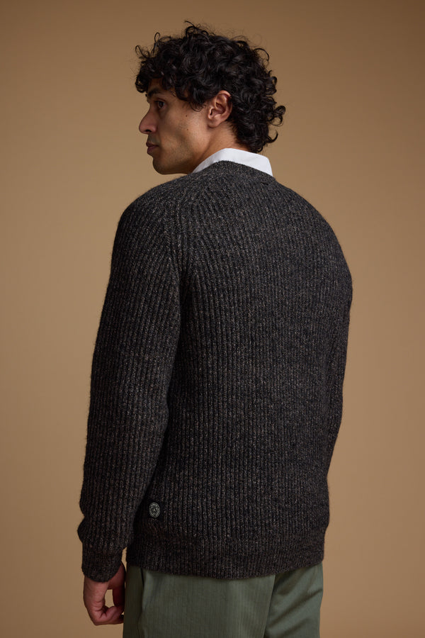 A man with curly dark hair stands sideways, facing backward, wearing the Barkers Hawthorn Woollen Crew sweater over a white shirt and green pants against a plain beige background.