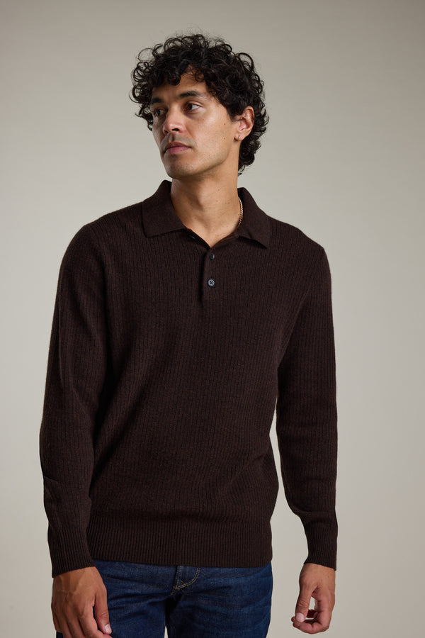 A man with curly dark hair wears the Barkers Braxton Merino Long Sleeve Polo in dark brown and blue jeans, standing against a plain light background and looking slightly to his left.
