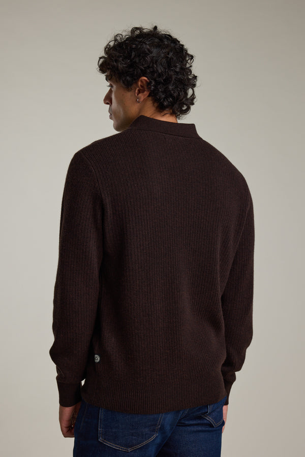 A person with short, curly dark hair faces away from the camera, wearing the Barkers Braxton Merino Long Sleeve Polo in dark brown and blue jeans against a plain light background.