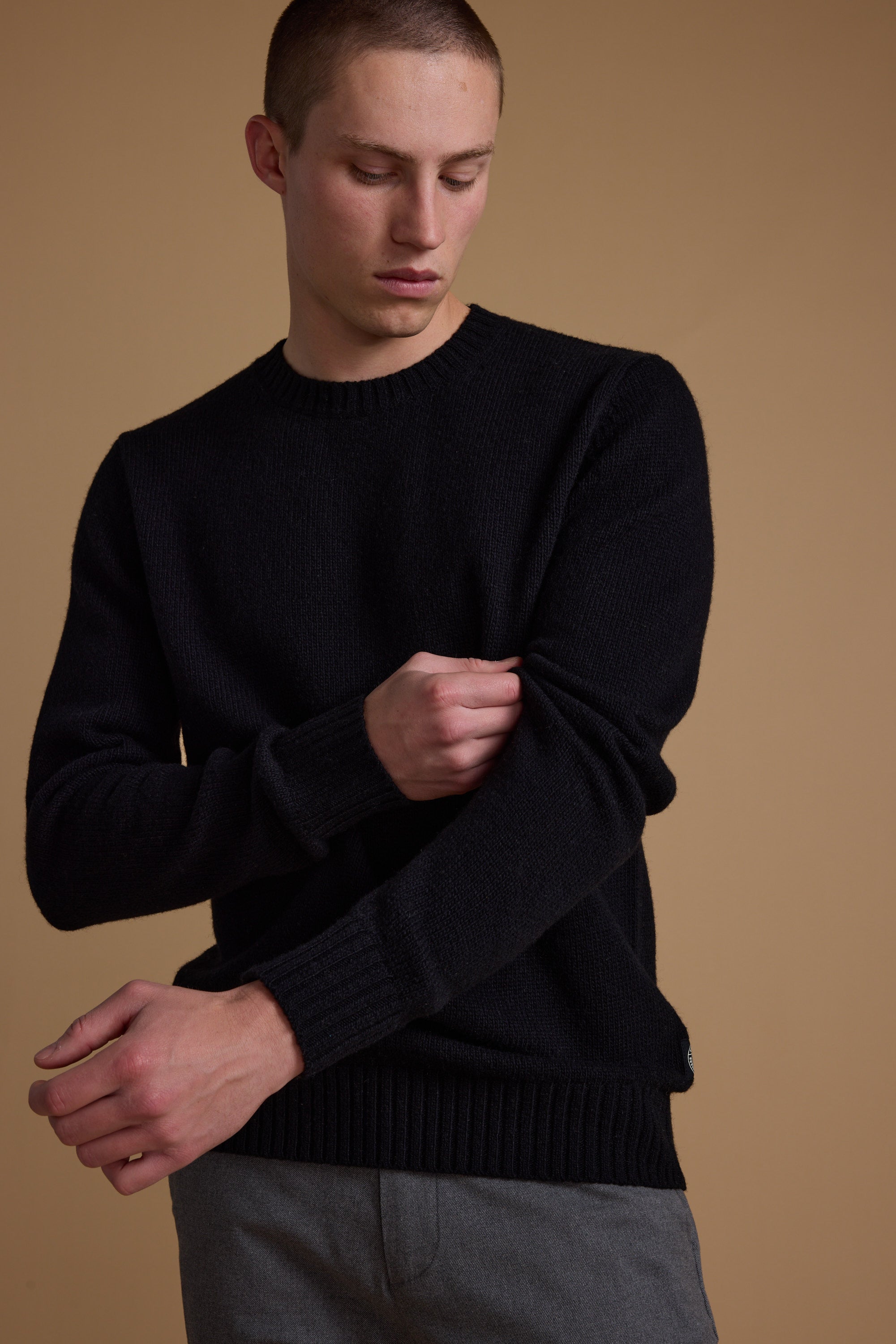A young man with short hair wears the Barkers Anglers Merino Crew in black and gray pants, standing against a plain beige background, adjusting his left sleeve while looking down.