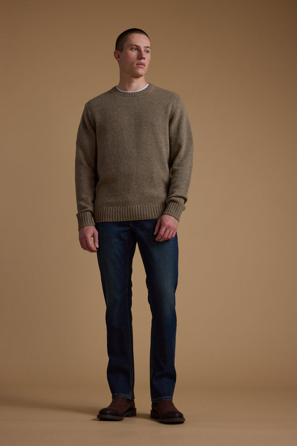 A man stands against a beige background wearing the Barkers Anglers Merino Crew sweater in brown, paired with blue jeans and brown shoes. He gazes slightly to the side with a neutral expression, his hands relaxed at his sides.