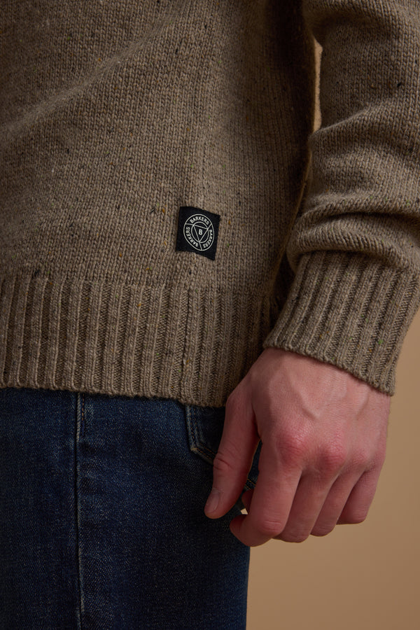 A close-up of a person wearing the Barkers Anglers Merino Crew—a brown fisherman’s knit sweater with a black and white circular logo tag—paired with dark blue jeans, highlighting the textured fabric and right hand.