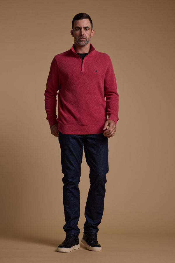 A man with short dark hair and a neutral expression faces forward against a beige background, wearing the Barkers Ketch Cotton 1/4 Zip red pullover with an embroidered motif, paired with dark blue jeans and black sneakers.