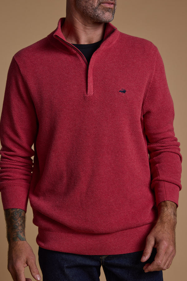 A man stands with his hands by his sides against a beige background, wearing the Barkers Ketch Cotton 1/4 Zip sweater in red, layered over a black shirt and paired with dark jeans.