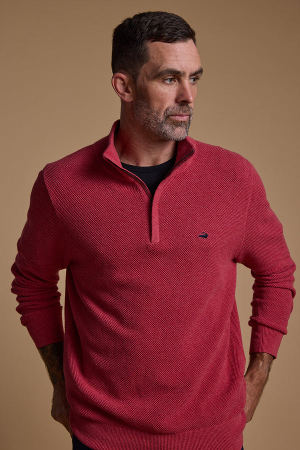 A man with short dark hair and a beard wears the Barkers Ketch Cotton 1/4 Zip in textured red over a black shirt, standing against a plain beige background, looking slightly left with his hands behind his back.