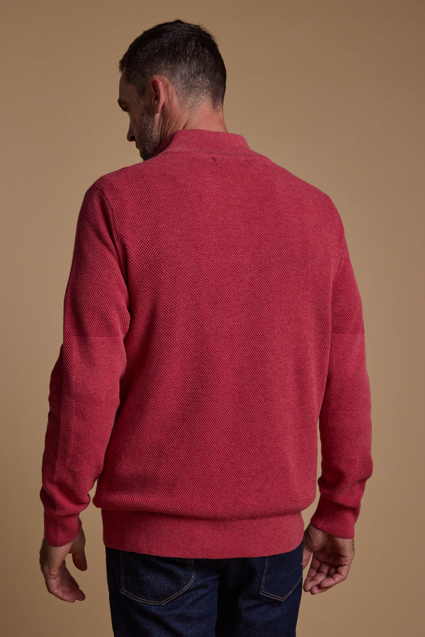 A man stands with his back to the camera, wearing the Barkers Ketch Cotton 1/4 Zip red sweater made from responsibly sourced cotton, paired with dark blue jeans, set against a plain beige background.