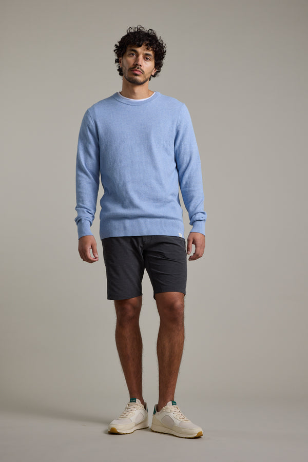 A man with short curly hair stands against a plain background, wearing the Barkers Dunes Texture Crew sweater in light blue, paired with dark gray shorts and white sneakers. He looks relaxed, arms resting at his sides.