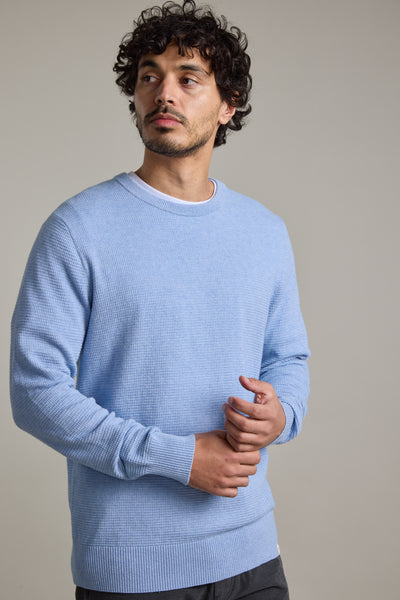 A man with curly hair and a short beard wears the Barkers Dunes Texture Crew in light blue and dark pants, standing against a plain light gray background, looking slightly to the side with a neutral expression.