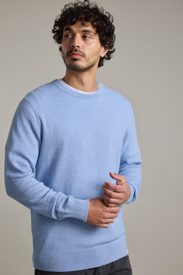 A man with curly hair and a short beard wears the Barkers Dunes Texture Crew in light blue and dark pants, standing against a plain light gray background, looking slightly to the side with a neutral expression.