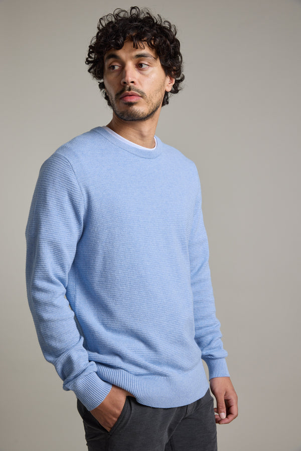 A man with curly hair and a slight beard models the Barkers Dunes Texture Crew, a light blue breathable sweater, paired with gray pants. He stands against a plain light background, gazing to the side with his hands in his pockets.