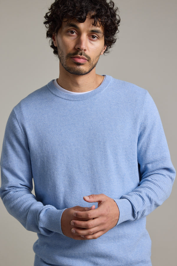 A man with curly dark hair and stubble wears the Barkers Dunes Texture Crew sweater in light blue, standing against a plain light gray background and looking directly at the camera with a neutral expression.