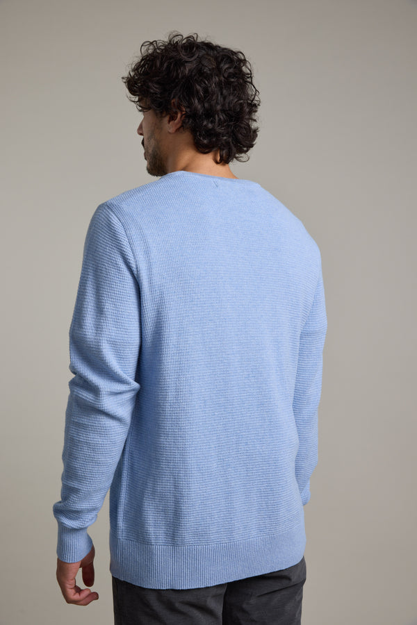 A man with curly dark hair stands with his back to the camera, wearing the Barkers Dunes Texture Crew in light blue and dark pants against a plain light background.