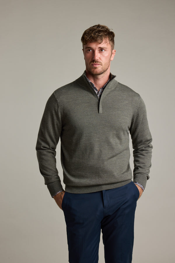 Man in Barkers Skyland ZQRX Merino 1/4 Zip and blue pants stands hands in pockets, plain backdrop.