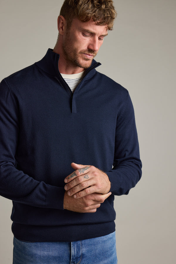Man in a navy Barkers Skyland ZQRX Merino 1/4 Zip and jeans looks down with hands clasped.