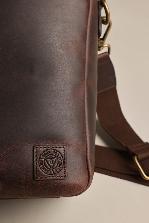 Close-up of the Barkers Wilkinson Leather Laptop Bag in dark brown, featuring a fabric shoulder strap, brass buckle, and an embossed circular logo patch on the lower corner.