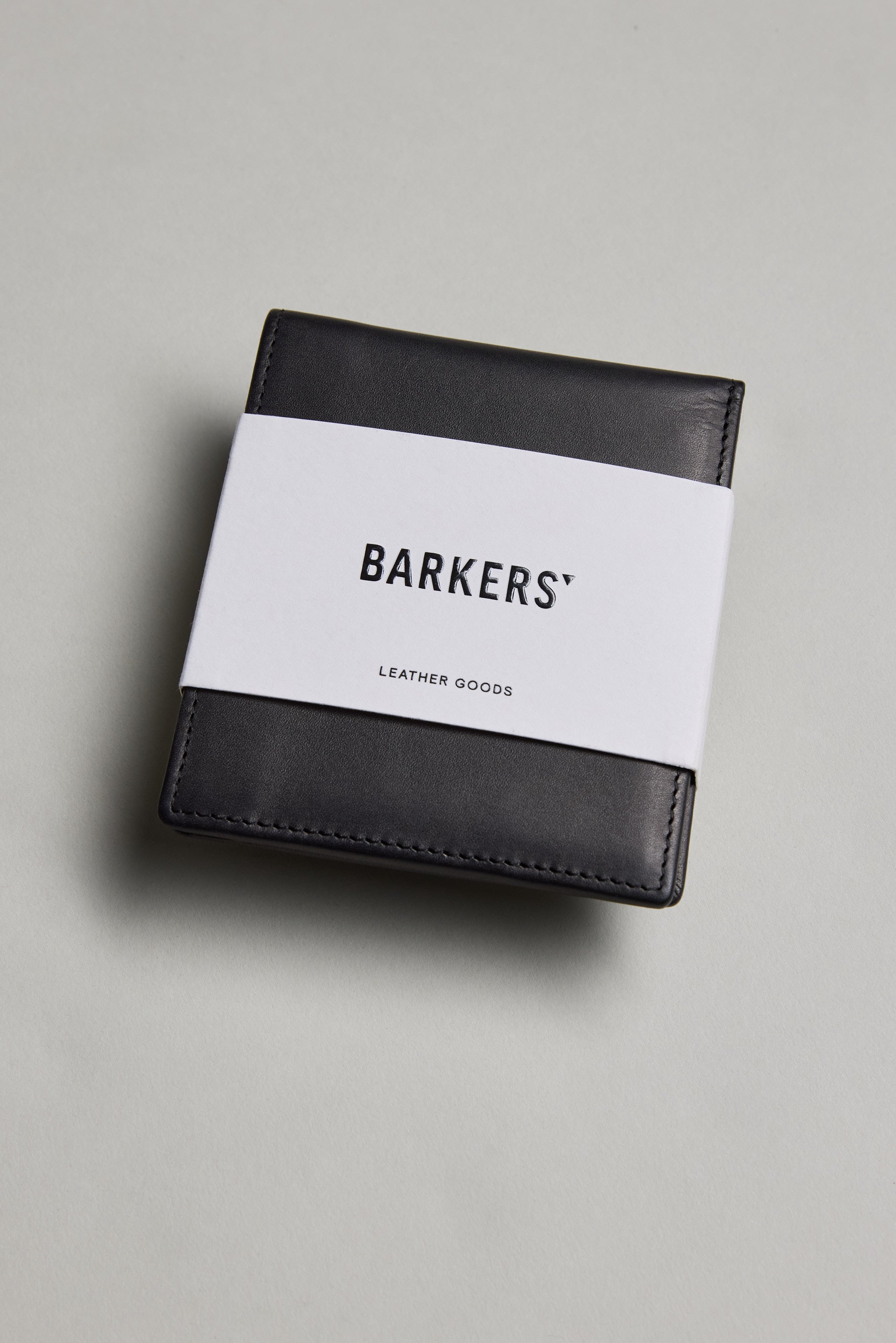 A black Harris Leather Fold Wallet by Barkers, wrapped in a white paper band with the brand name, is placed on a light gray surface.