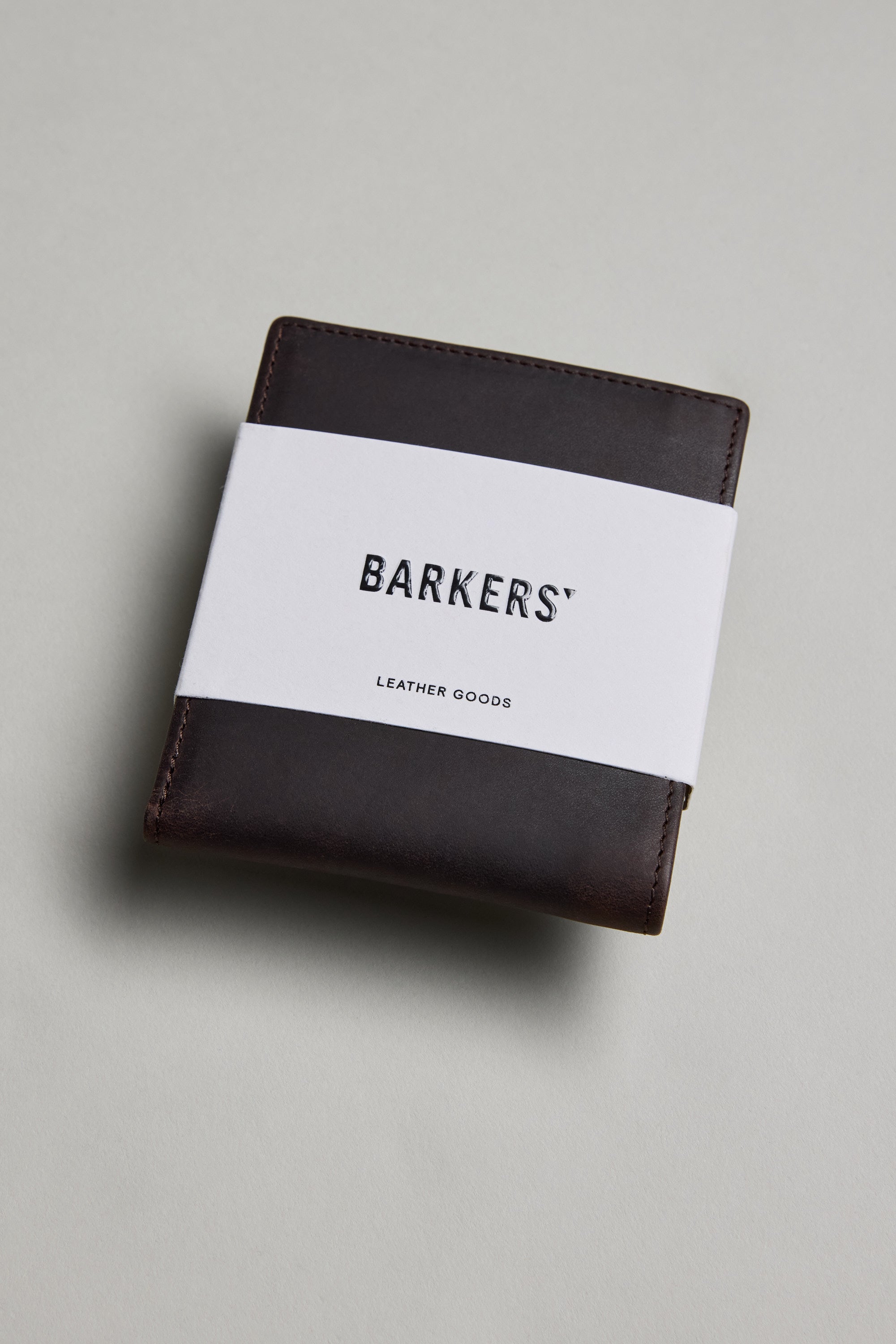 A dark brown Harris Leather Fold Wallet by Barkers, wrapped with a white paper band labeled BARKERS LEATHER GOODS, rests on a light gray surface.