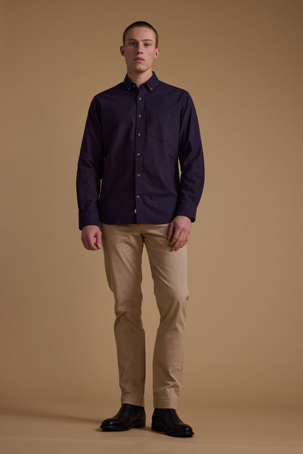 A young man stands against a tan background, wearing the Barkers Thatcher Oxford Shirt in dark purple crafted from responsibly sourced cotton, beige pants, and black shoes. He faces forward neutrally with his arms at his sides.