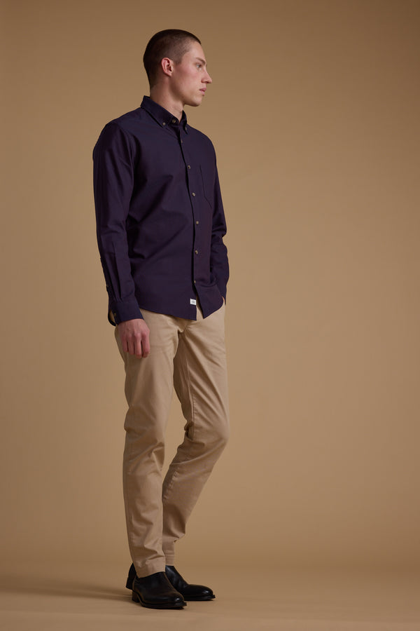 A person stands against a plain brown background, wearing the Barkers Thatcher Oxford Shirt in dark purple, made from responsibly sourced cotton, paired with beige pants and black shoes, left hand in pocket and looking to the side.