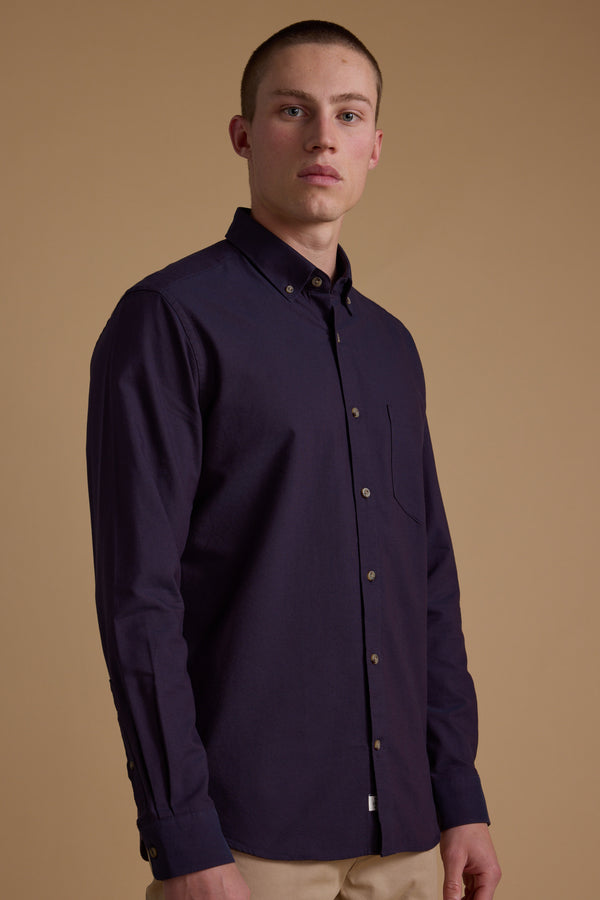 A person stands against a beige background, wearing the Barkers Thatcher Oxford Shirt in dark purple with a tailored fit and beige pants, looking directly at the camera with a neutral expression.