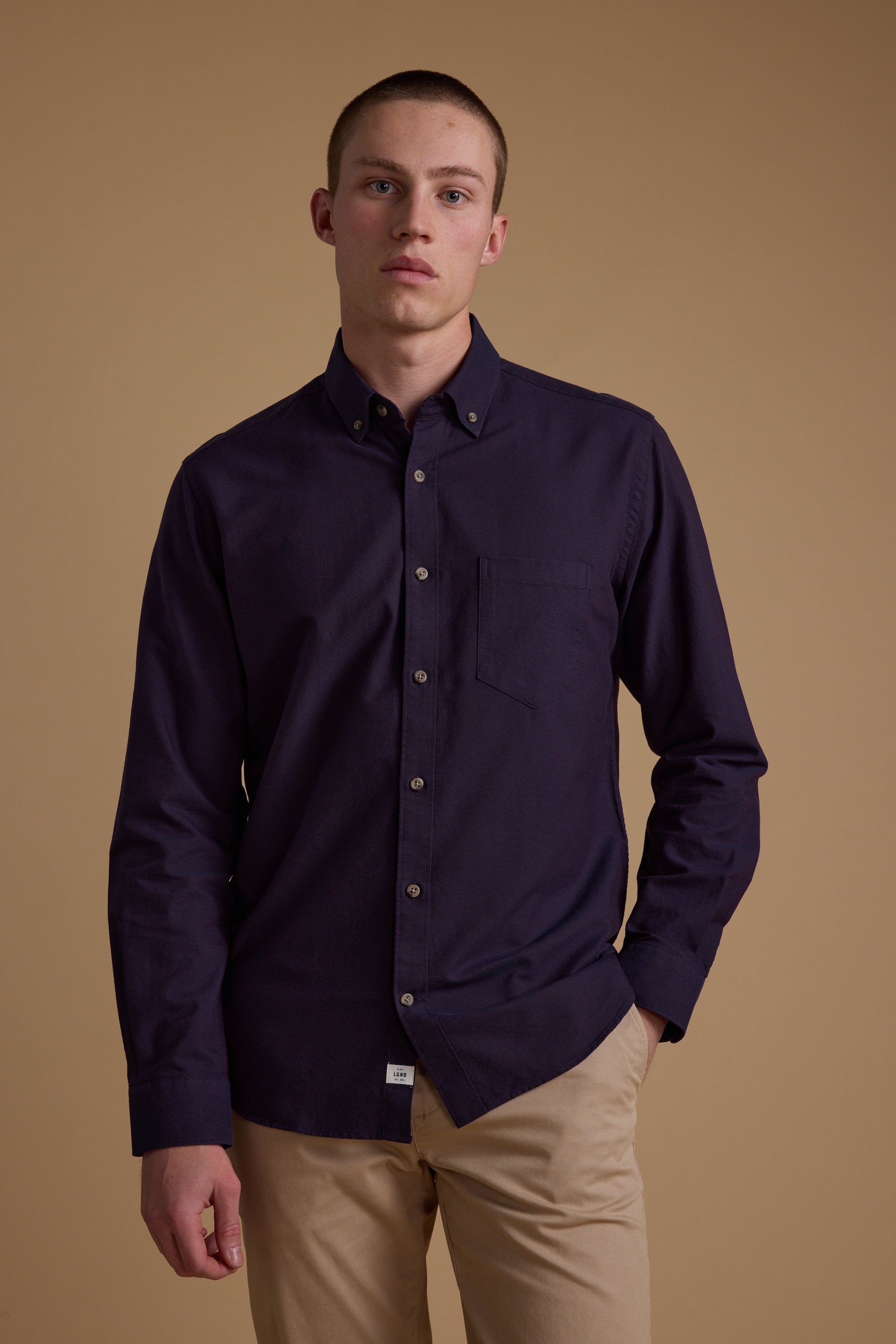 A young man stands against a beige background, wearing the Barkers Thatcher Oxford Shirt in dark purple and light khaki pants, with one hand in his pocket and a neutral expression. The shirt is tailored fit and made from responsibly sourced cotton.