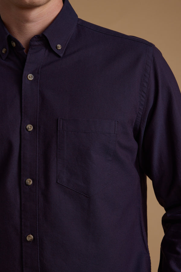 A person wearing the Barkers Thatcher Oxford Shirt in dark purple, featuring a front pocket and tailored fit, is photographed from shoulders to waist against a plain light brown background.
