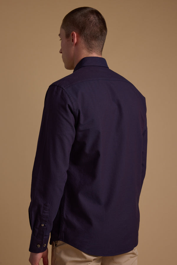 A person with short hair stands with their back to the camera, wearing Barkers’ Thatcher Oxford Shirt in dark purple, crafted from responsibly sourced cotton, paired with light-colored pants against a plain beige background.