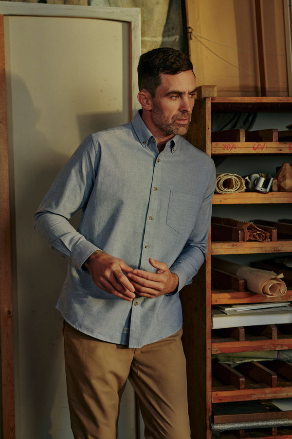 A man wearing the Barkers Thatcher Oxford Shirt in light blue, made from responsibly sourced cotton, and khaki pants stands indoors by wooden shelves stocked with tools and materials, gazing thoughtfully to the side.
