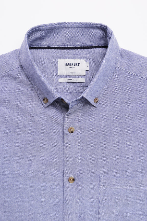 A neatly folded light blue Barkers Thatcher Oxford Shirt with a collar, chest pocket, and inside brand label. Made from responsibly sourced cotton, this tailored-fit shirt has brown buttons and is laid flat on a white background.