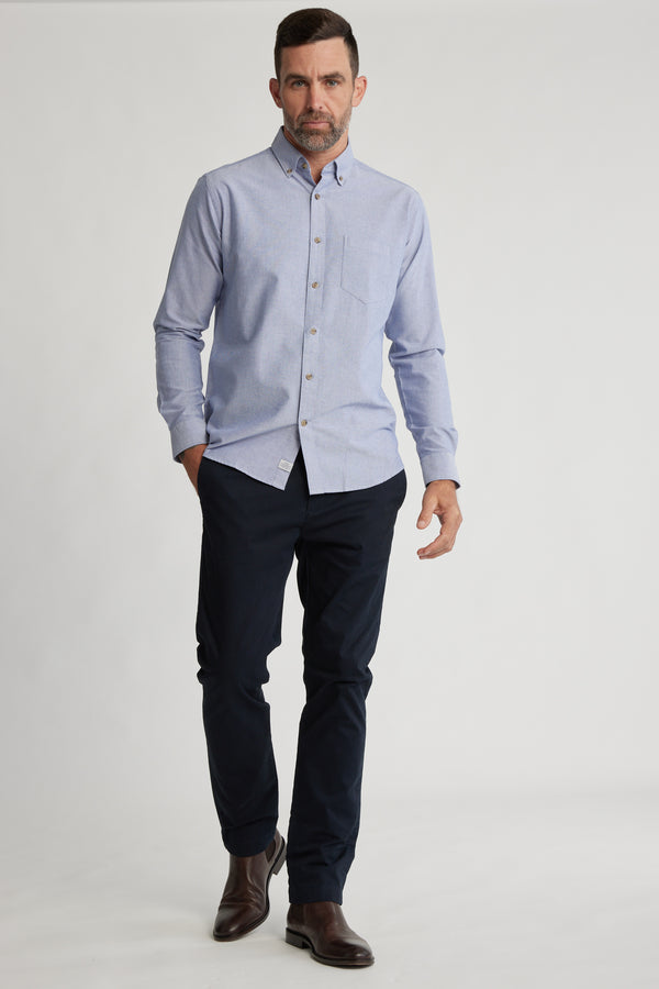 A man wearing the Barkers Thatcher Oxford Shirt in light blue, paired with dark navy trousers and brown boots, walks forward against a plain white background.