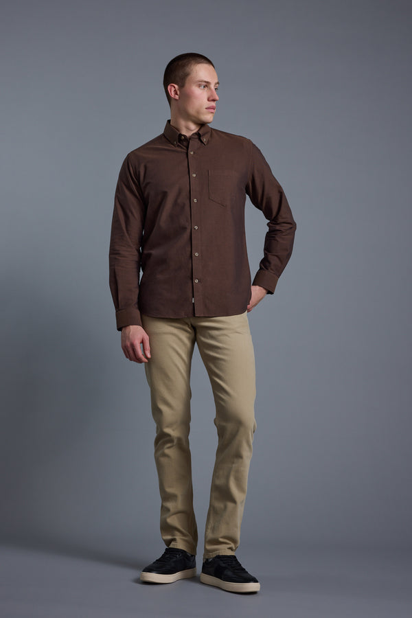 A man stands against a gray background, wearing the Barkers Thatcher Oxford Shirt in brown, beige tailored-fit pants, and black sneakers. His left hand is in his pocket as he looks to his left.