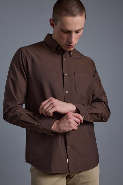Wearing the Barkers Thatcher Oxford Shirt in brown and beige pants, a person looks down as they fasten a cuff button, set against a plain gray background.