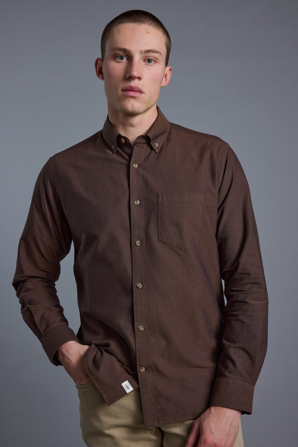 A man with short hair wears the Barkers Thatcher Oxford Shirt in brown, made from responsibly sourced cotton, paired with beige pants. He stands against a plain gray background, one hand in his pocket and a neutral expression.