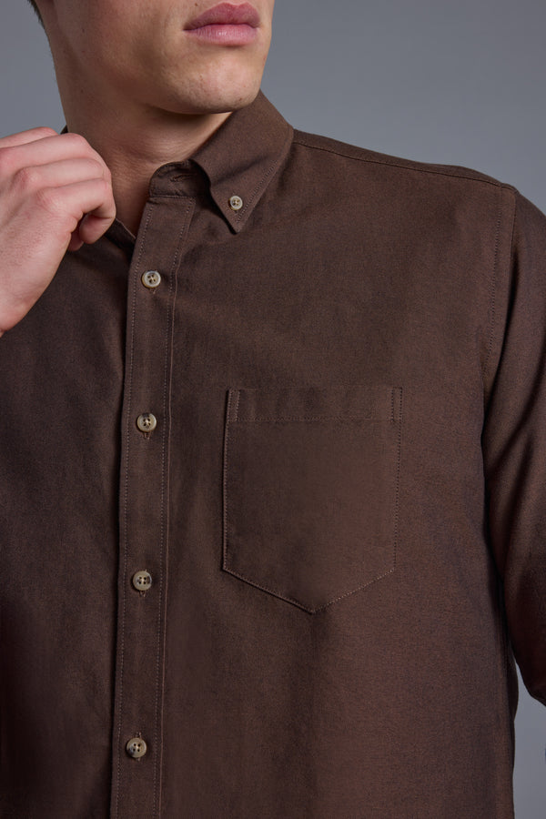A person holds the collar of a Barkers Thatcher Oxford Shirt in brown, featuring a front pocket and tailored fit, shown from shoulders to mid-torso against a plain gray background.