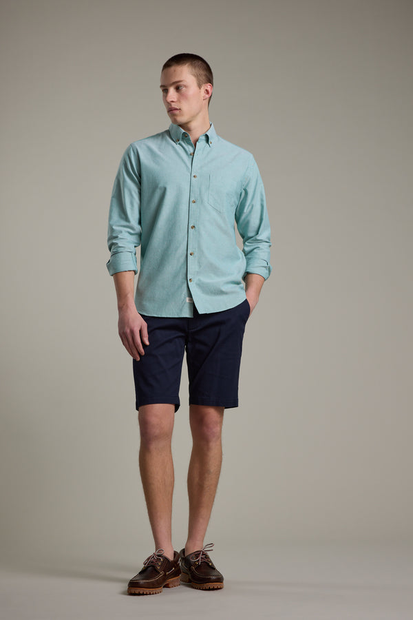 A man stands against a plain background wearing the Barkers Thatcher Oxford Shirt in light blue with sleeves rolled up, dark navy shorts, and brown boat shoes. The tailored shirt is crafted from responsibly sourced cotton. He looks to his left, left hand in pocket.